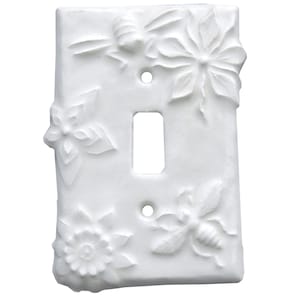 Könnte beinhalten: Weiße Keramik-Lichtschalterabdeckung mit geprägten Blumen- und Bienenmotiven. Die Abdeckung hat eine rechteckige Öffnung für den Schalter und zwei Schraubenlöcher. Die Reliefs zeigen Blumen und Bienen.