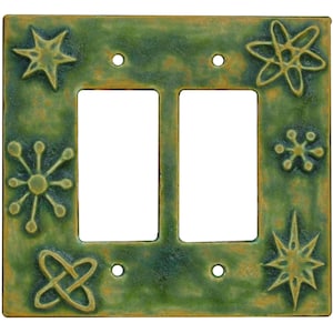 May include: A double light switch wall plate with a mottled green and brown finish. The rectangular plate features two vertical cutouts for switches and four screw holes. Embossed starburst and atomic-style geometric shapes decorate the corners.