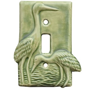 以下が含まれることがあります： 2羽のサギがレリーフで描かれた、明るい緑色のセラミック製スイッチカバー。1羽は立っており、もう1羽は水に向かって曲がっています。カバーは長方形で、スイッチ用の穴と2つのネジ穴があります。