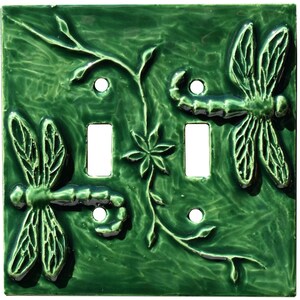 Könnte beinhalten: Eine quadratische, glänzend dunkelgrüne Keramik-Doppellichtschalterabdeckung mit einem erhabenen Reliefdesign aus zwei Libellen und einer zentralen Blume mit rankenartigen Stielen. Die Platte hat vier Schraublöcher und zwei rechteckige Öffnungen.