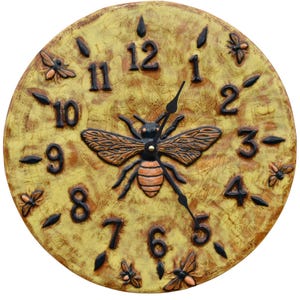 May include: A circular decorative clock with a bee motif, in shades of yellow and brown. The clock face displays black numerals, black hands, and a central bee design. Additional bee and flower details are included.