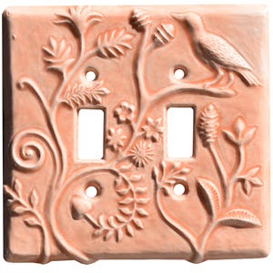 May include: A peach-colored, ceramic double light switch cover with a raised relief design. The design features a bird perched on a branch, along with floral and foliate elements. The cover has two rectangular switch openings and three screw holes.