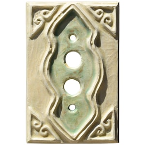 Może przedstawiać: Prostokątna ceramiczna osłona włącznika światła z kremowym tłem i centralnym, jasnozielonym, wydłużonym wzorem z trzema okrągłymi otworami. Krawędzie zdobione są dekoracyjnymi zawijasami.