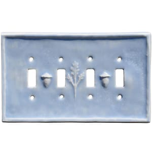 May include: A pale blue ceramic four-gang light switch plate with a glossy finish. It features a raised leaf and acorn design. The plate has four rectangular switch openings and four small mounting holes.