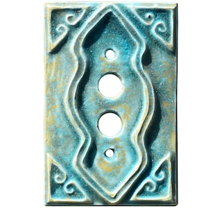 Może przedstawiać: Prostokątna, turkusowa osłona włącznika światła z dekoracyjnym, wypukłym wzorem. Posiada trzy okrągłe otwory i bogaty, symetryczny wzór ze złotymi akcentami. Ogólna estetyka jest vintage lub antyczna.