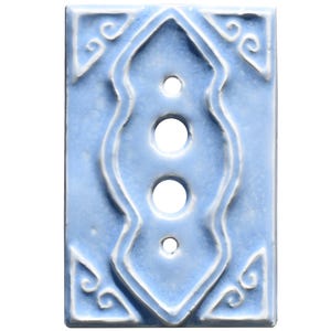 Może przedstawiać: Jasnoniebieska ceramiczna płytka przełącznika z dekoracyjnym wzorem. Płytka ma trzy okrągłe otwory i podniesiony, ozdobny wzór pośrodku. Narożniki zdobią detale w kształcie zawijasów, tworząc estetykę vintage.