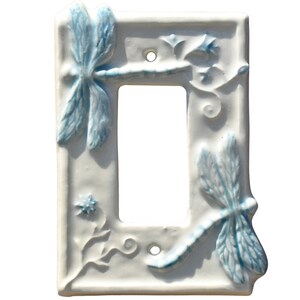 Puede incluir: Cubierta de interruptor de luz de cerámica blanca con diseños de libélulas y flores azules en relieve. La cubierta rectangular presenta dos libélulas, una en la parte superior y otra en la inferior, con una abertura central.