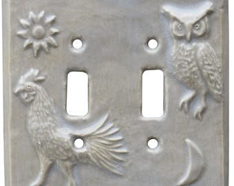 May include: A light gray double light switch cover with embossed designs. The cover features a sun, a rooster, an owl, and a crescent moon. The switch plate has two rectangular openings for the switches and two circular holes for the screws.