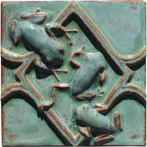 Op de afbeelding: Vierkante keramische tegel met drie blauwgroene kikkers en oranje accenten. De kikkers zijn geplaatst in een geometrisch patroon op een gevlekte groene achtergrond. Rustieke, handgemaakte uitstraling.