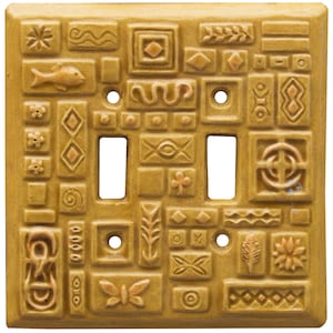 May include: A mustard yellow double light switch wall plate featuring a raised relief pattern of various geometric shapes, a fish, a butterfly, flowers, and abstract symbols across the entire rectangular surface.