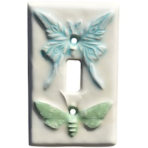 May include: A white ceramic light switch cover featuring two embossed butterfly designs. The top butterfly is light blue, and the bottom butterfly is green. The cover has a single-switch opening.