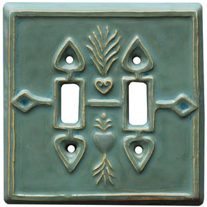 May include: Square, teal ceramic double light switch cover with a raised, decorative design. The design includes two rectangular switch openings, heart and diamond shapes, and stylized plant motifs. The edges and design details are highlighted with a gold color.