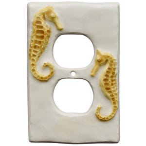 May include: A white ceramic double outlet cover featuring two raised seahorse designs in shades of tan and gold. The seahorses are positioned on either side of the outlet openings, adding a coastal or nautical theme to the decor.