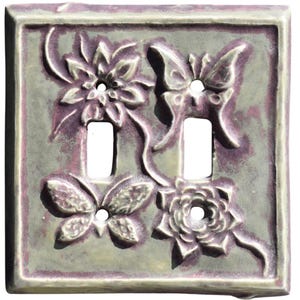 Puede incluir: Una cubierta decorativa para interruptor de luz doble con un diseño floral y de mariposas. La cubierta cuadrada tiene un fondo verde claro y flores y mariposas moradas en relieve. Los bordes son de un verde más claro.