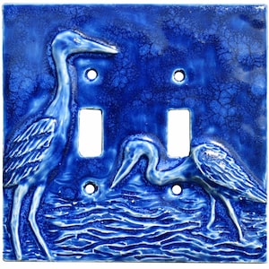 Peut inclure: Une plaque de recouvrement pour double interrupteur carrée en émail bleu cobalt profond avec une finition craquelée. La plaque présente un relief en relief de deux échassiers à long cou dans l'eau, avec deux découpes rectangulaires.