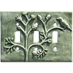 May include: A green ceramic light switch cover with a raised design of a tree branch with leaves, flowers, and a bird. The switch cover has three toggle switches.