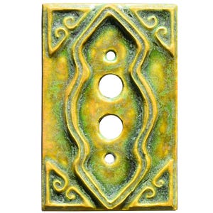 Może przedstawiać: Prostokątna osłona włącznika światła w stylu antycznym z postarzanym wykończeniem. Osłona posiada centralny, wydłużony wzór z trzema okrągłymi otworami i dekoracyjnymi zawijasami w rogach. Kolorystyka to połączenie złota i zieleni.