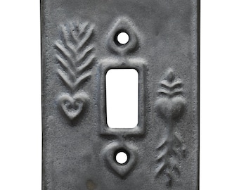 May include: A rectangular, dark gray metal light switch cover with a central rectangular opening and two circular holes. The cover features embossed floral and heart-shaped designs on either side of the switch opening. The metal has a textured, aged appearance.
