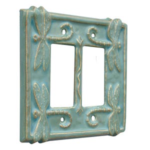 Może przedstawiać: Podw&oacute;jny włącznik światła w kolorze jasnoniebieskim z wzorem ważki. Pokrywa ma złotą ramkę i jest wykonana z ceramiki.