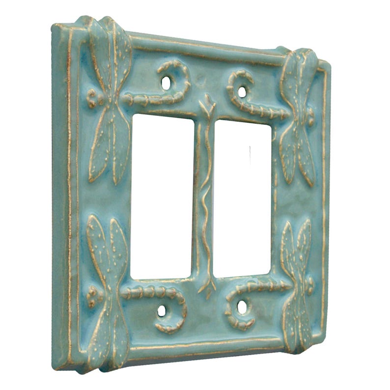 Może przedstawiać: Podw&oacute;jny włącznik światła w kolorze jasnoniebieskim z wzorem ważki. Pokrywa ma złotą ramkę i jest wykonana z ceramiki.