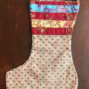May include: A handmade Christmas stocking with a patchwork design. The top section features horizontal stripes of red, light blue, and yellow patterned fabric. The lower portion is a cream-colored fabric with a repeating paisley pattern and stitched details.