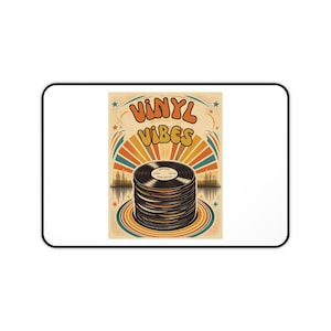 May include: A rectangular desk mat featuring a graphic illustration of a stack of black vinyl records. The text VINYL VIBES appears in orange and yellow bubble letters above the records, set against a background of sunburst rays and stars.