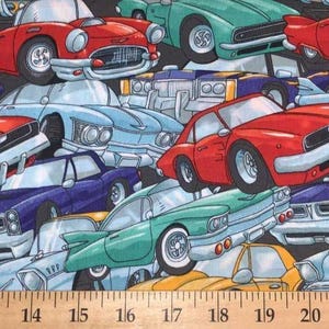 May include: A fabric print featuring a repeating pattern of cartoon-style vintage cars in red, teal, light blue, purple, and yellow on a black background, positioned above a wooden ruler showing measurements from 10 to 24 inches.