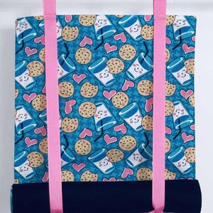 May include: A fabric roller blind with a blue background featuring a pattern of smiling milk cartons, cookies, and pink hearts. The blind has a black bottom and is suspended by pink ribbons.