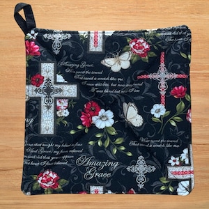 May include: A square quilted potholder with a black background featuring white ornate crosses, red and white flowers, butterflies, and the text Amazing Grace along with lyrics from the hymn. It includes a black fabric loop for hanging.