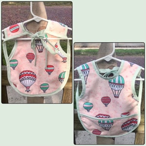 May include: Two pink and green baby bibs with a hot air balloon print. The bibs have a tie closure and a pocket on the front.