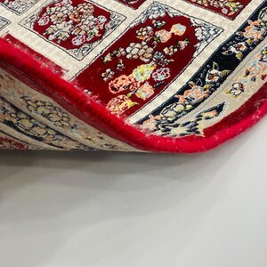 May include: A section of a red oriental rug with a floral pattern. The rug has a cream-colored border and a red fringe. The design features floral motifs in various colors, including red, yellow, and blue. The rug is partially rolled up, revealing its underside.