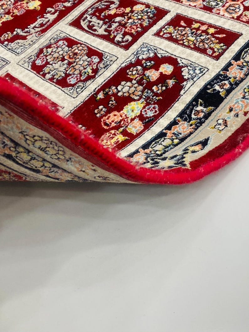 May include: A section of a red oriental rug with a floral pattern. The rug has a cream-colored border and a red fringe. The design features floral motifs in various colors, including red, yellow, and blue. The rug is partially rolled up, revealing its underside.