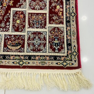 May include: A section of a red oriental rug with a cream-colored fringe. The rug features a repeating pattern of floral designs in red, cream, and other colors. The border is black with floral accents. The fringe is cream-colored.