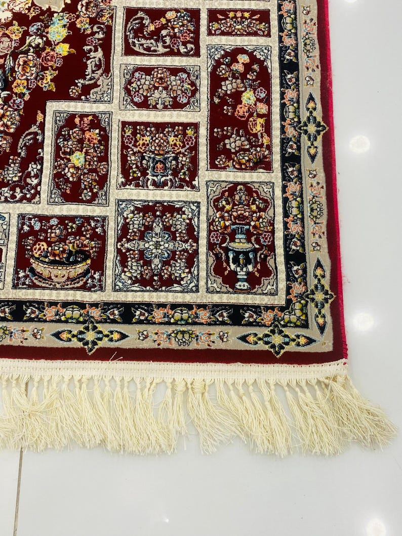 May include: A section of a red oriental rug with a cream-colored fringe. The rug features a repeating pattern of floral designs in red, cream, and other colors. The border is black with floral accents. The fringe is cream-colored.