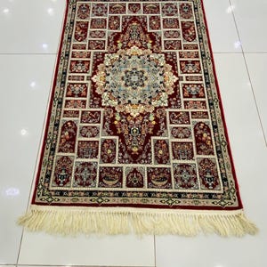 May include: A rectangular red rug with an intricate floral pattern and a cream-colored fringe. The rug features a central medallion design surrounded by smaller, repeating motifs in shades of red, cream, and blue. The rug's dimensions are not visible.
