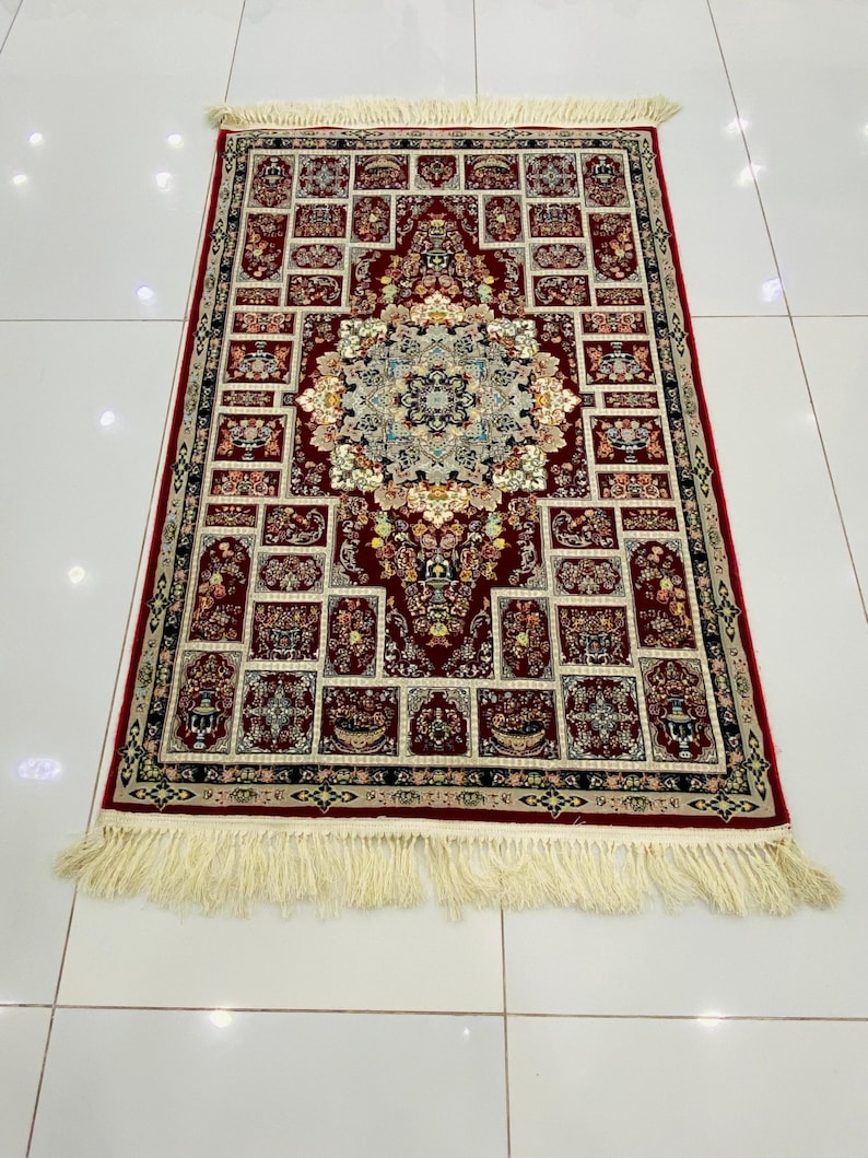 May include: A rectangular red rug with an intricate floral pattern and a cream-colored fringe. The rug features a central medallion design surrounded by smaller, repeating motifs in shades of red, cream, and blue. The rug's dimensions are not visible.