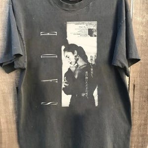 Puede incluir: Camiseta gris oscuro con una imagen en blanco y negro de una mujer con pendientes de aro. La palabra "SADE" está impresa verticalmente a la izquierda. Texto japonés impreso a la derecha. La camiseta se muestra en una percha de madera.