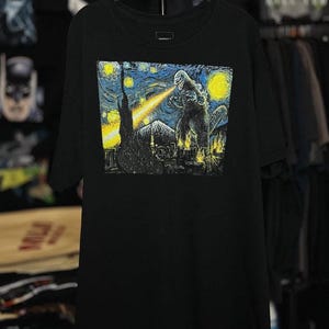 Può includere: T-shirt nero con una grafica di Godzilla in una scena notturna stellata. Il design include un raggio giallo che emana dalla bocca di Godzilla, sullo sfondo di un cielo blu e giallo vorticoso e uno skyline.