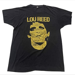 Puede incluir: Camiseta negra con un gráfico amarillo del rostro de Lou Reed y el texto "LOU REED" encima de la imagen. La camiseta es de estilo cuello redondo clásico, probablemente de algodón o tejido similar, y parece vintage.