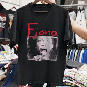 Może przedstawiać: Czarny t-shirt z czerwono-białą grafiką piosenkarki Fiony Apple. Grafika przedstawia czarno-biały obraz Fiony Apple ze słowem "Fiona" w kolorze czerwonym nad jej wizerunkiem. Koszulka wisi na drewnianym wieszaku.