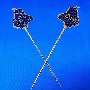 May include: Two silver-toned pins crossed against a blue background. Each pin features a blue map-shaped design with white text. The pins are likely decorative or functional, possibly for clothing or accessories.