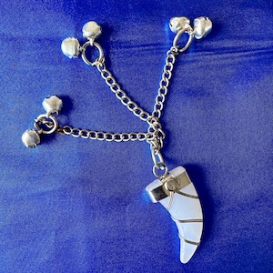 Puede incluir: Un collar de cadena de plata con un colgante blanco en forma de diente y varias campanillas de plata. El colgante está envuelto con alambre de plata.