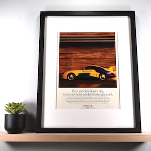 May include: Framed vintage Toyota race car advertisement. The image features a yellow, blue, and green race car speeding across a blurred background. The text reads, "For a preview of race day, turn your head quickly from right to left."