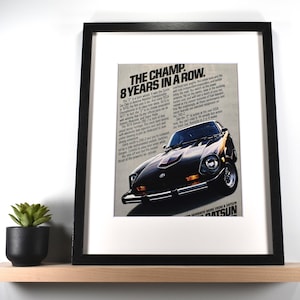 May include: Framed print featuring a vintage Datsun car advertisement. The black framed print has a white mat and a black car with the text "THE CHAMP. 8 YEARS IN A ROW." A small potted succulent sits on a wooden shelf below the print.
