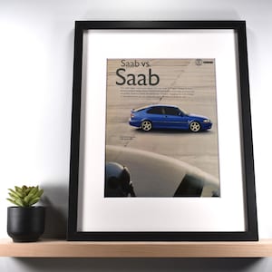 May include: A framed vintage advertisement print featuring a blue Saab car on a runway with the text Saab vs. Saab. The print is housed in a black frame with a white mat. The advertisement includes a small Saab logo and descriptive text.