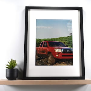 May include: Framed print featuring a red pickup truck driving on a dirt road. The truck has a silver grill and headlights. The print is in a black frame with a white mat, displayed on a wooden shelf with a small potted plant.