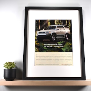May include: Framed print featuring a silver SUV in a forest setting. The print includes the text "THE ANSWERING MACHINE FOR THE CALL OF THE WILD." The Toyota 4Runner is the focus of the image, set against a backdrop of trees.