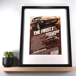 May include: Framed print featuring a vintage advertisement for the Datsun 280-ZX. The poster has a black frame and white matting. The ad text reads "THE FIRST ZX" and "A CREATURE OF COMFORT". A small potted plant sits on a wooden shelf below.