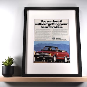 May include: Framed vintage advertisement for a red Saab convertible. The ad features the text "You can love it without getting your heart broken." A small potted succulent sits on a wooden shelf below the framed image.