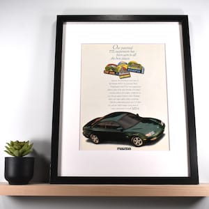 May include: Framed advertisement for a green Mazda MX-6 car. The ad features the car, text, and logos. The frame is black, and the image is displayed on a light-colored shelf with a small potted plant.